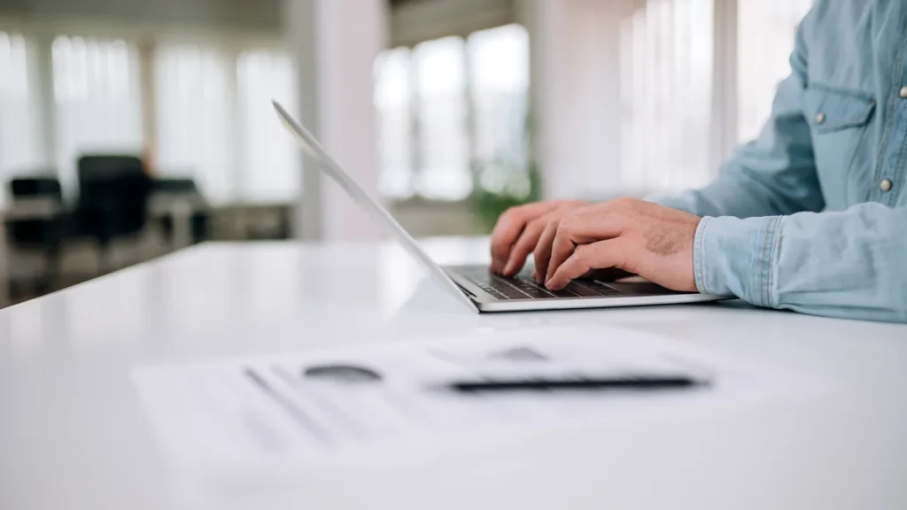 Mains tapant sur un ordinateur portable dans un bureau lumineux, avec des documents posés sur la table